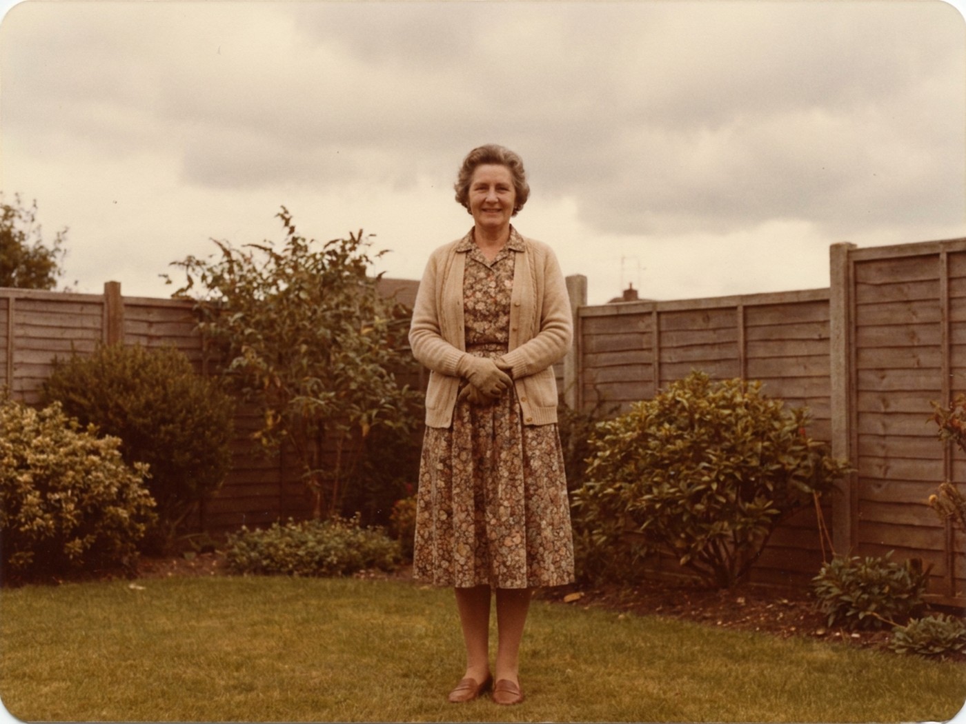 A colour photograph in the style of 1984: a woman standing alone in a garden, in a long sleeved floral dress and cardigan.