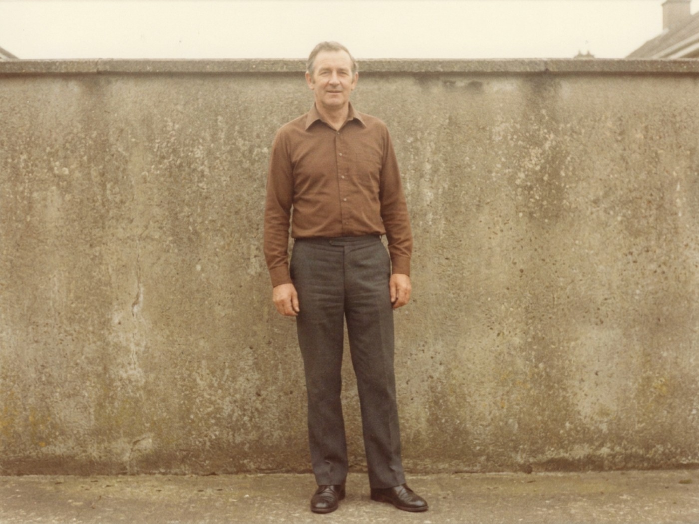 A colour photograph in the style of 1984: a man standing alone in front of a garden wall, in a long sleeved shirt and dark trousers.