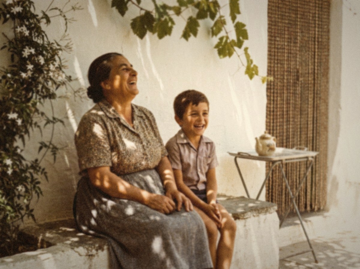 A photograph from 1978 of a grandmother laughing on a stone bench in a courtyard, with her young grandson sitting beside her.